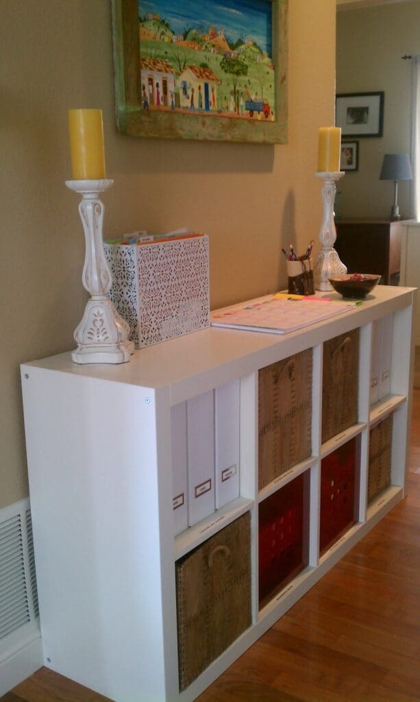 A white shelving unit with storage bins, folders, and baskets sits against a wall; two yellow candles on decorative holders, a metal organizer, and art supplies are on top. Above, a colorful painting completes this cheerful Mommy Central.