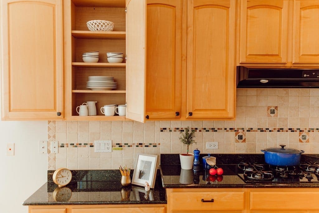 A well-organized kitchen with wooden cabinets, one open to reveal neatly stacked plates and mugs. The black countertop features a clock, a plant, utensils, onions, a framed picture, and a blue pot on the stove. The beige backsplash is adorned with decorative tiles for added charm.