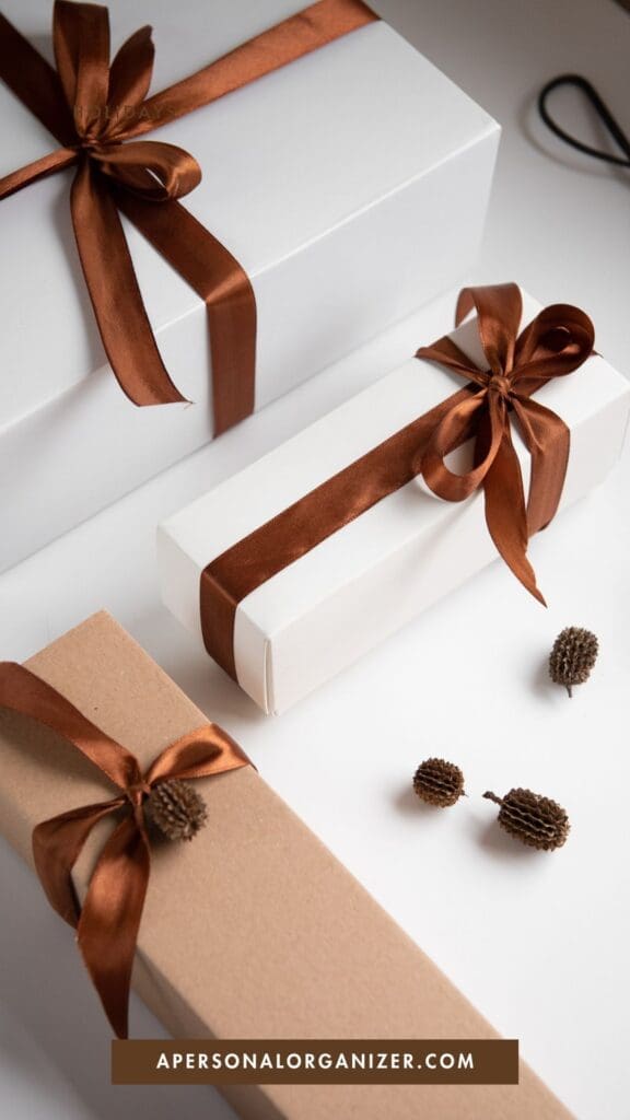 Three gift boxes, two white and one brown, are decorated with shiny brown ribbons—perfect for holiday planning. Small decorative pinecones are scattered beside the boxes. A website label reads “apersonalorganizer.com” at the bottom.