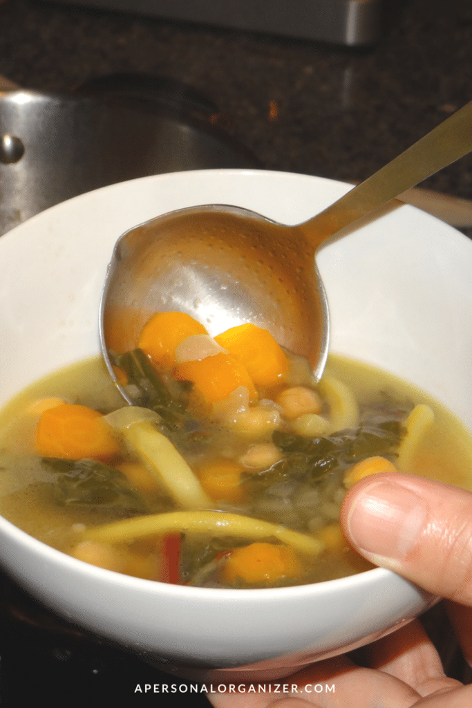 A hand holding a white bowl of vegetable soup containing carrots, beans, greens, and noodles, with a ladle hovering above, filled with more soup. A stove and pot are visible in the background—perfect for recovering from Thanksgiving. "apersonalorganizer.com" is written at the bottom of the image.