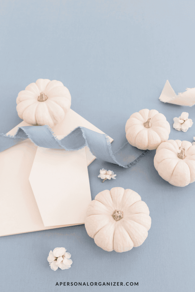 A flat lay of small white pumpkins arranged on a light blue background with pale pink envelopes and a light blue fabric ribbon invites you to give thanks. White flowers are scattered among the pumpkins and envelopes. The website "apersonalorganizer.com" is displayed at the bottom.