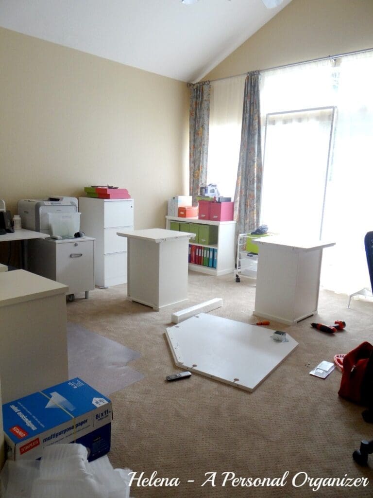 A room in the process of being organized, with disassembled white furniture on the floor, boxes, and office supplies scattered around. There are file cabinets and shelves against the wall, perfect for home office organization ideas. Sunlight streams through a large window with floral curtains.