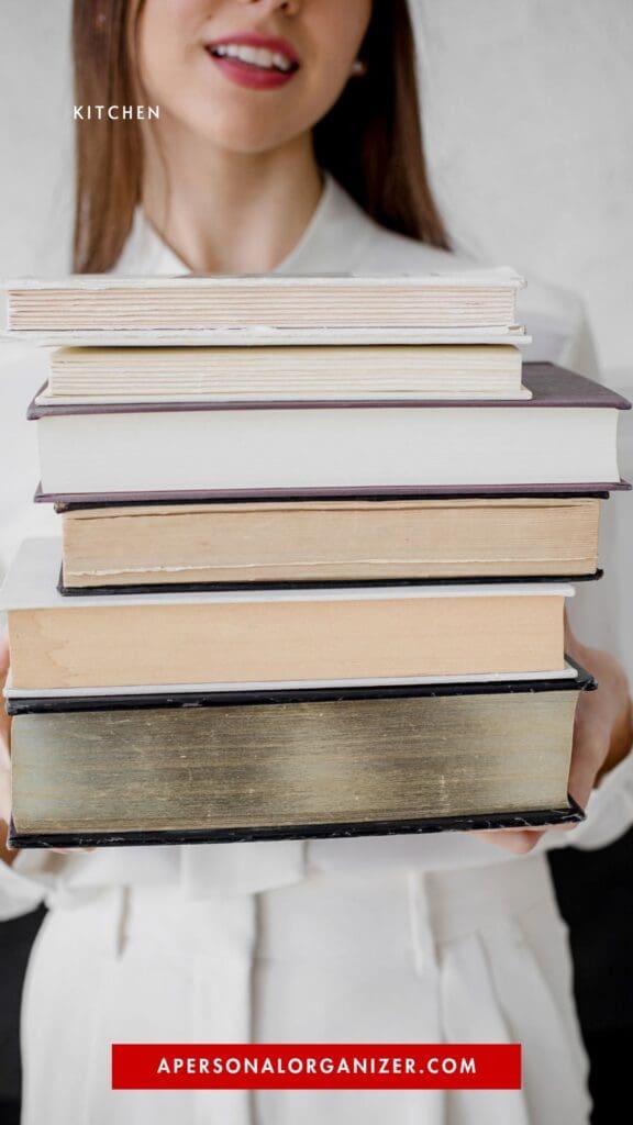 A woman in a white outfit holds a tall stack of books, symbolizing declutter and organization. The word "KITCHEN" appears near her face, while a red bar at the bottom reads "APERSONALORGANIZER.COM".