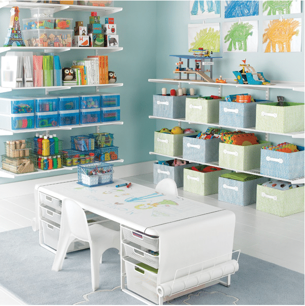 A neatly organized, clutter-free children's playroom with white shelves holding books, toys, and art supplies. Green and blue storage bins contain various toys. A white table with drawings and two white chairs is in the center, and colorful children's artwork is displayed on the wall.