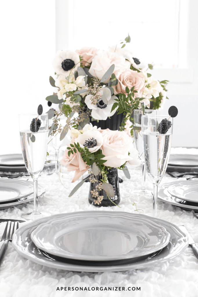 Elegant fall table decorations feature a centerpiece of white and blush roses with greenery. Four place settings with grey plates and black cutlery surround the arrangement. Each place includes a champagne flute with a small black decorative detail inside, adding a touch of autumnal charm.