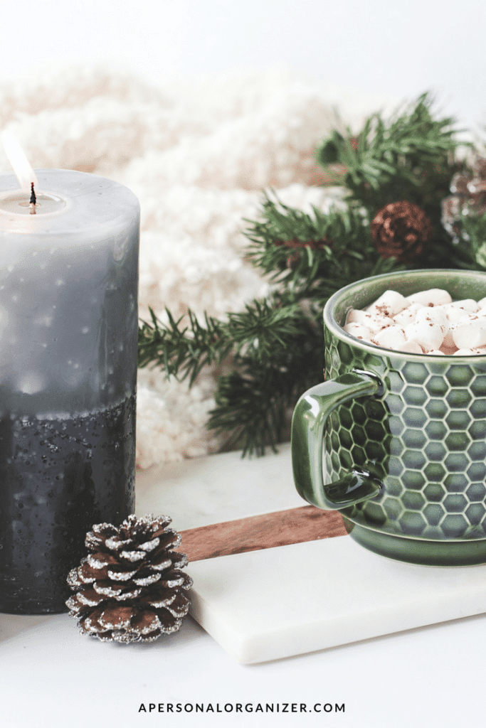 A cozy scene features a green mug with a honeycomb pattern filled with marshmallow-topped hot chocolate. The mug sits on a white tray next to a pine cone and a lit gray candle, perfect for some holiday organizing in the background alongside evergreen branches and a fluffy white blanket.