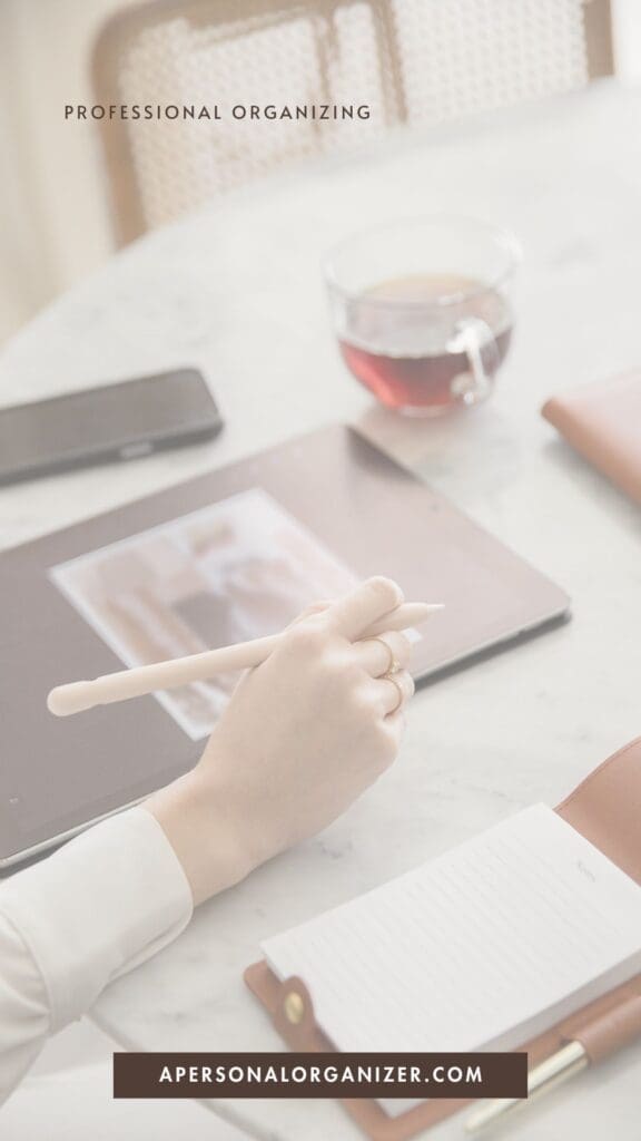 A person writes in a notebook beside a tablet showing a photo, a cup of tea, and a phone on a light desk—perfect for declutter inspiration. Text reads "Professional Organizing" and "apersonalorganizer.com".