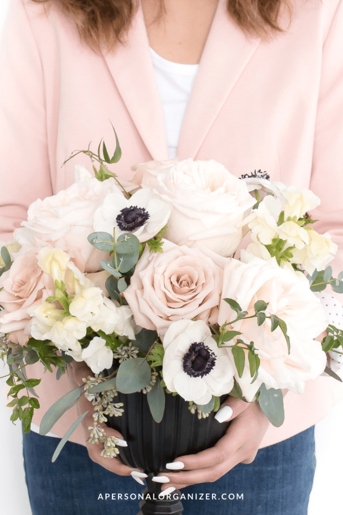 A person in a light pink blazer, preparing for hosting, holds a bouquet of flowers featuring pink roses, white anemones with dark centers, and eucalyptus leaves in a black vase.