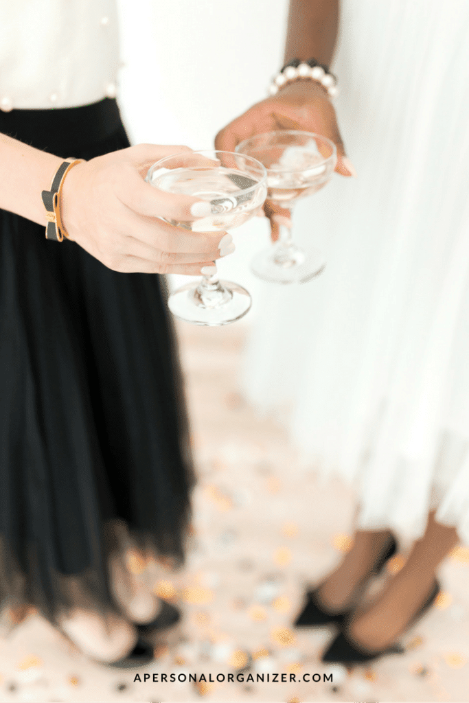 Two individuals, dressed in elegant black and white attire, hold champagne glasses while standing on a confetti-strewn floor. One wears a black skirt, and the other a white skirt. Amidst the festive atmosphere, they chat about their New Year's resolutions. The website "apersonalorganizer.com" is visible at the bottom.