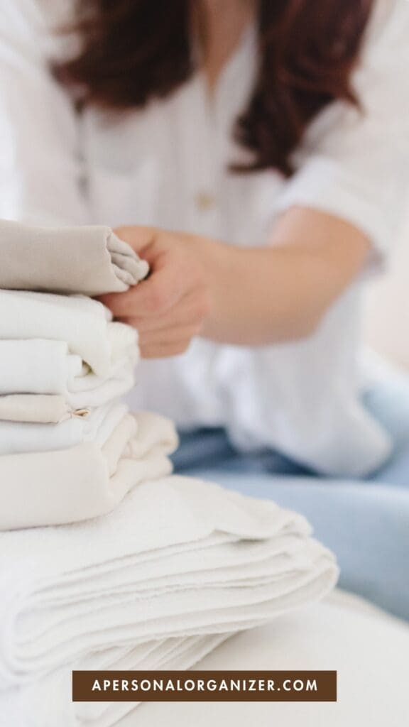 A person in a white shirt folding a stack of neatly arranged white and beige linens, demonstrating how to fold fitted sheets, with the text "APERSONALORGANIZER.COM" at the bottom.