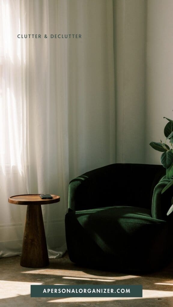 A sunlit room with a dark green armchair, a small round wooden side table, sheer white curtains, and a leafy plant creates an inviting space for declutter and organizing. Text reads "Clutter & Declutter" and "apersonalorganizer.com".