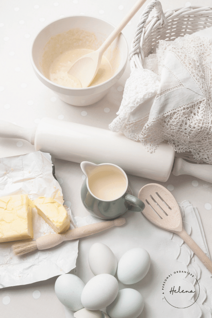 Amidst a soft, vintage ambiance, baking magic unfolds with eggs, butter on foil, and a cup of cream at the ready. A bowl of batter awaits the wooden spoon's embrace. Nearby, a Baking Ingredients Organizer showcases essentials including a rolling pin and spatula beside a cloth-lined wicker basket.