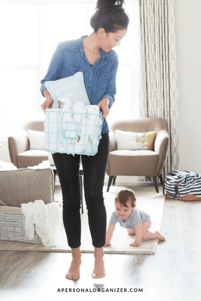 A woman in a denim shirt and black pants holds a laundry basket, looking down at a baby crawling on the floor. The room features light-colored furniture and decor, creating a cozy atmosphere that highlights effective home management. The text "apersonalorganizer.com" is visible at the bottom of the image.