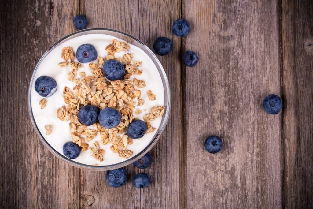 A bowl of yogurt topped with granola and fresh blueberries sits on a rustic wooden surface, perfectly organized for an appetizing display. Additional blueberries are scattered around the bowl on the wooden planks.