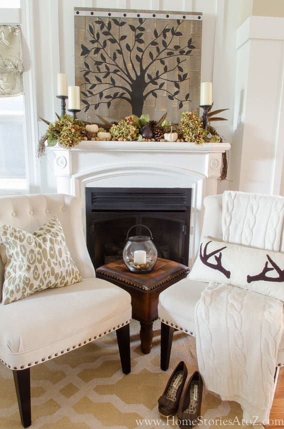 Cozy living room with a fireplace adorned with candles and fall garland, perfect for showcasing fall mantel ideas. Two cream chairs surround a small wooden table with a glass lantern. A white knit blanket with deer antlers drapes over one chair. Geometric rug on the floor.