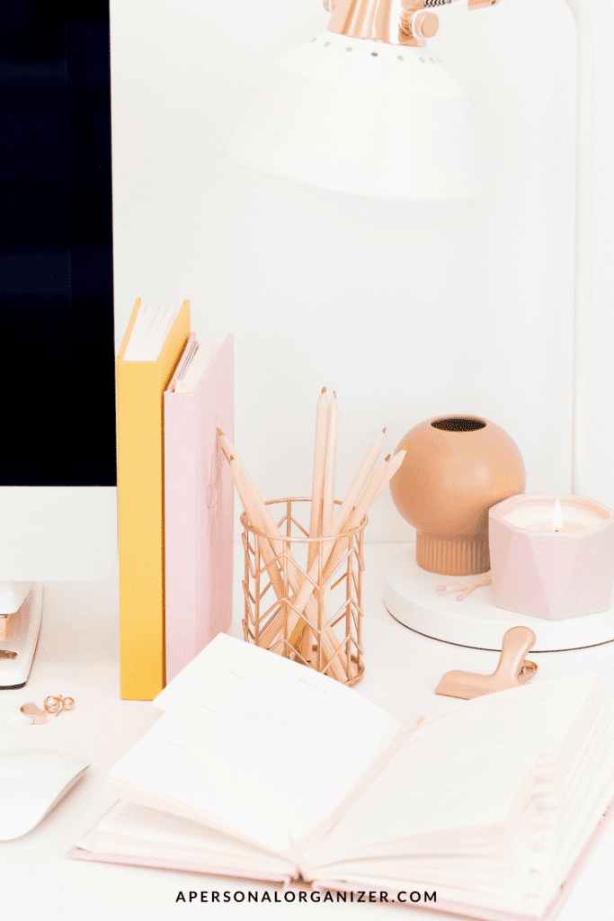 A neatly organized white desk featuring an open notebook, a set of pencils in a holder, two pastel-colored closed journals, a small round vase, a lit candle, and an angled white desk lamp. A portion of a computer screen, perhaps displaying email inbox organization tips, is visible on the left side.