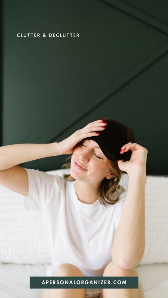 A woman in a white shirt sits on a bed, smiling with her eyes closed and adjusting a black sleep mask on her head—embracing organization. Text reads "Clutter & Declutter" and "apersonalorganizer.com.