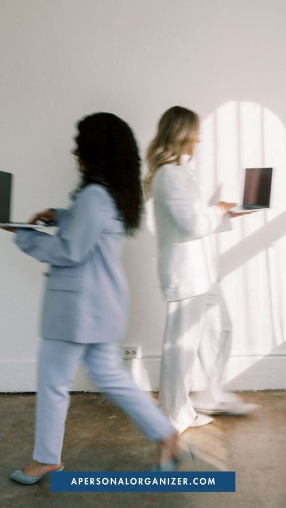 Two women in business attire walk past each other with open laptops, blurred in motion against a white wall. Sunlight casts shadows as they stride purposefully—declutter your workflow today. A website link appears at the bottom.