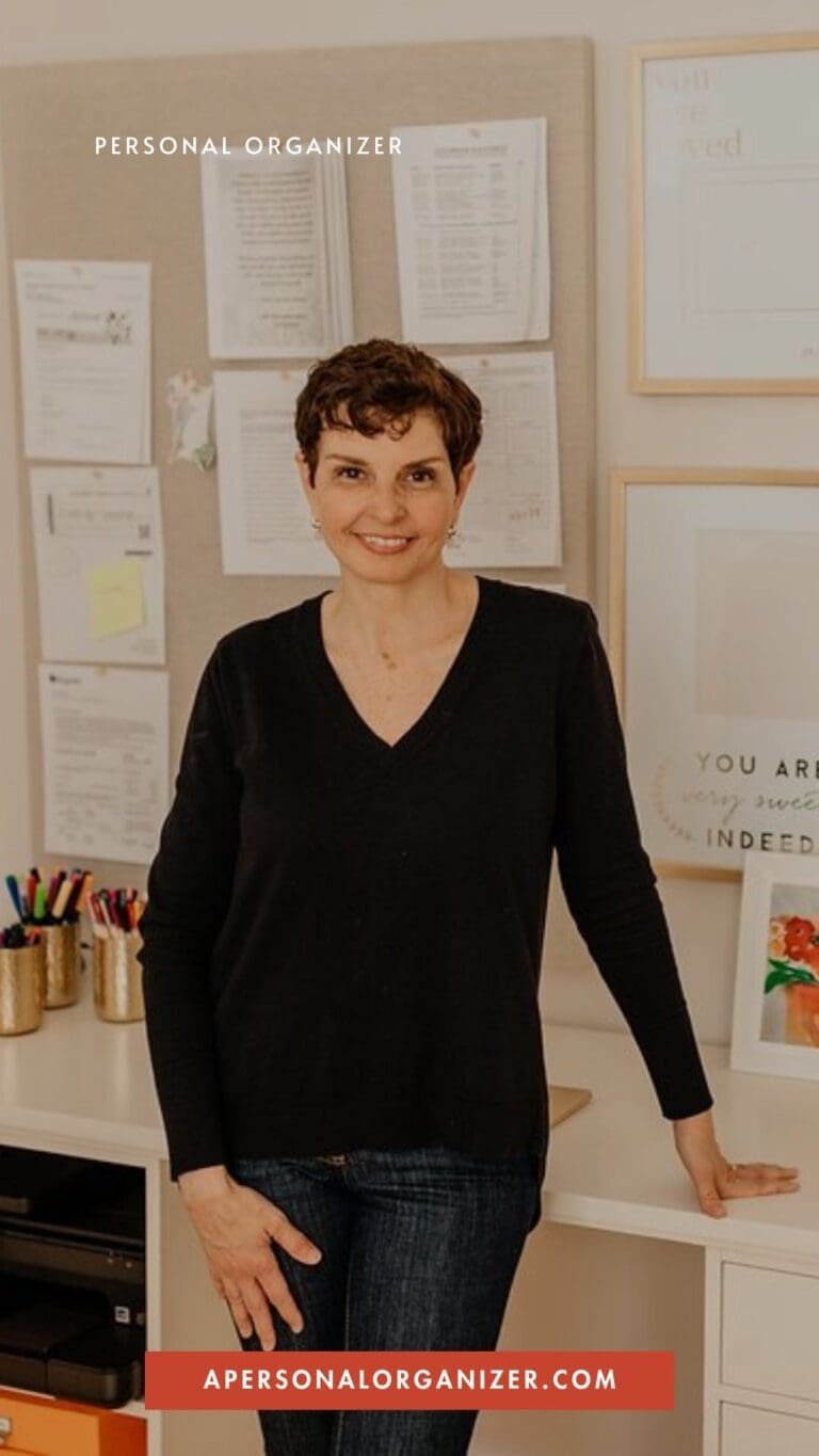 A woman with short curly hair, wearing a black sweater and jeans, stands smiling in an organized office space with papers pinned to a board behind her. Text reads "Personal Organizer" and "apersonalorganizer.com." She’s passionate about declutter and organizing.