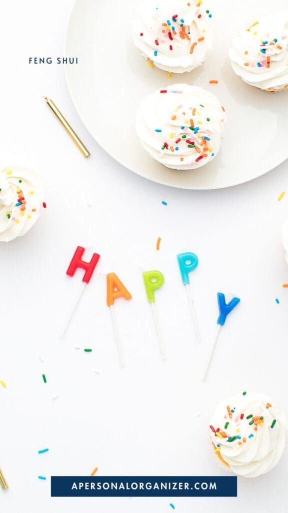 Cupcakes with white frosting and rainbow sprinkles surround colorful candles spelling “HAPPY” on a tidy white background. A gold pen and sprinkled crumbs are also visible. The text reads "Feng Shui" and "apersonalorganizer.com".
