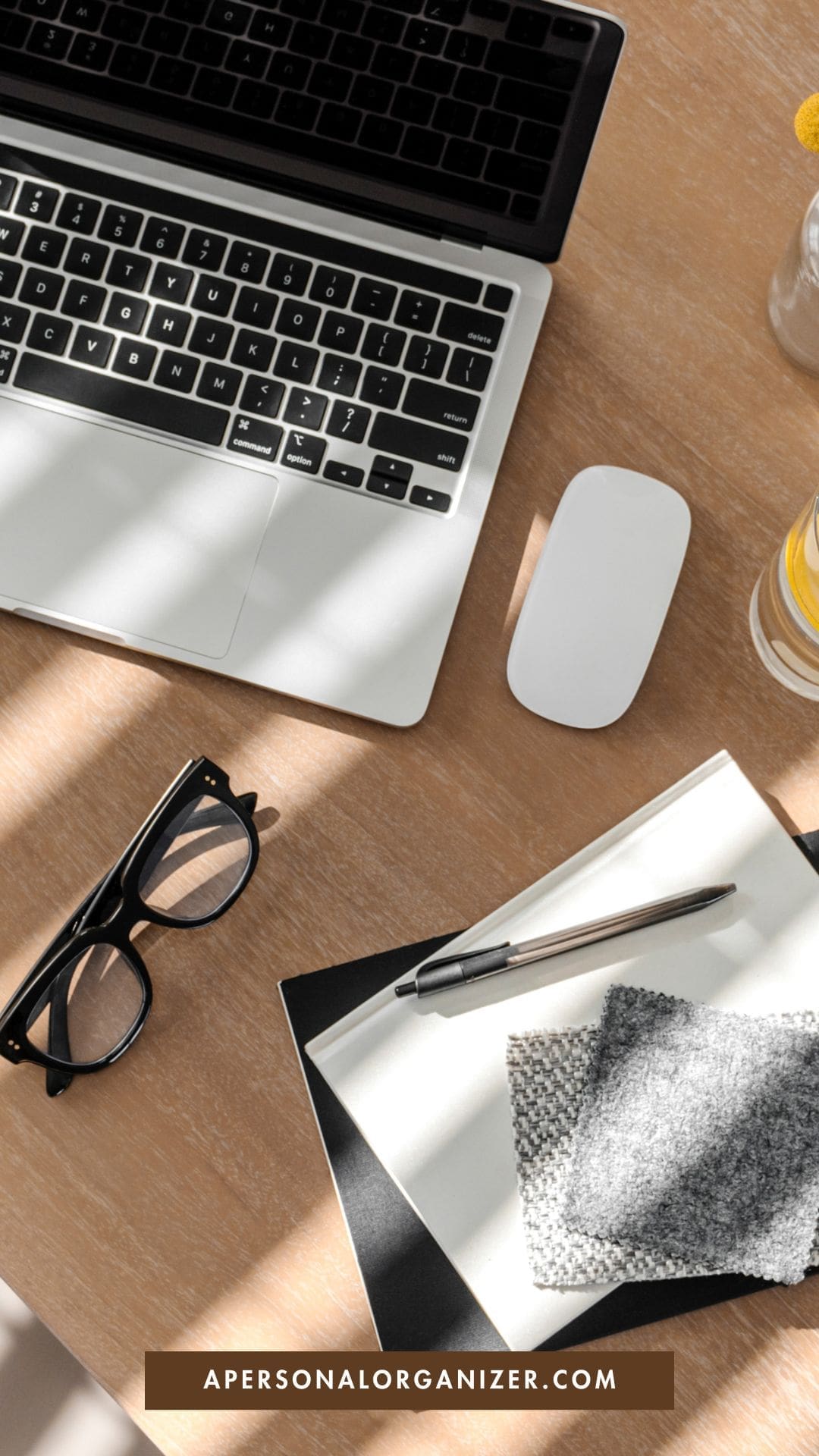 A desk with a laptop, wireless mouse, black eyeglasses, a pen on an open notebook, fabric swatches, and a glass of water with lemon—perfect for any personal organizer. The text "APERSONALORGANIZER.COM" is displayed at the bottom.