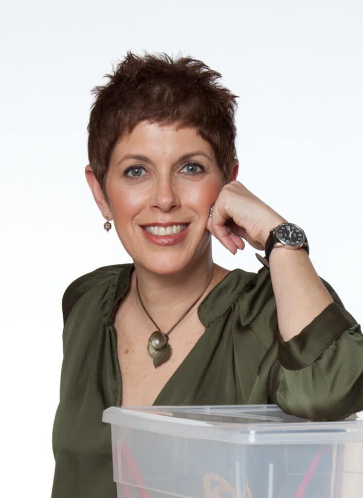 A smiling woman with short brown hair, wearing a green blouse, watch, and pendant necklace, rests her elbow on a clear plastic storage box against a white background—ready to get organized.