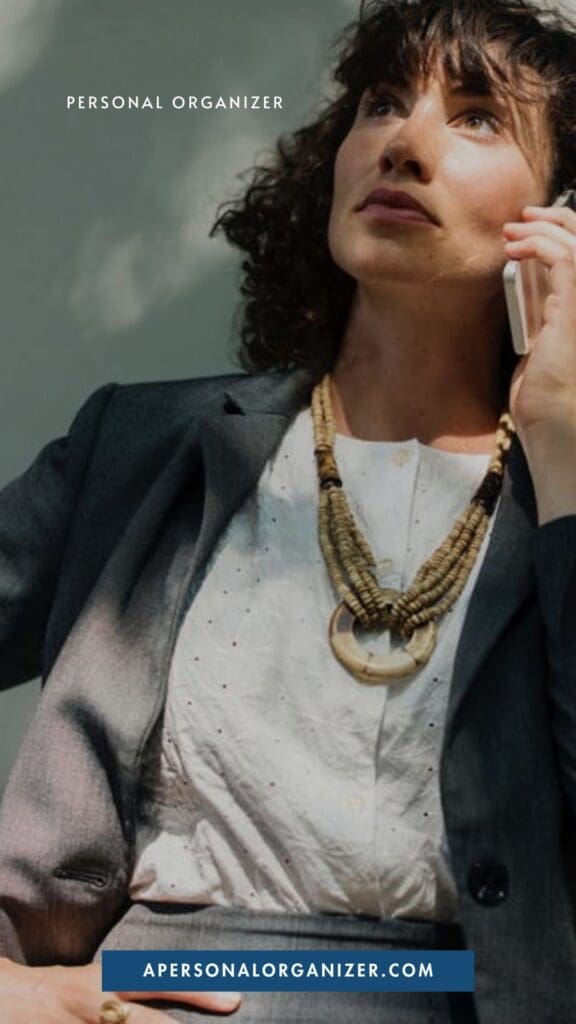 A woman in a business suit talks on her phone outdoors, ready to help you declutter your life. She wears a white blouse, gray blazer, and chunky necklace. Text reads "Personal Organizer" and "apersonalorganizer.com" at the bottom.