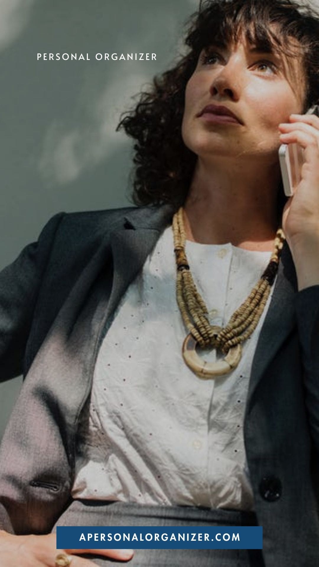 A woman in a business suit talks on her phone outdoors, ready to help you declutter your life. She wears a white blouse, gray blazer, and chunky necklace. Text reads "Personal Organizer" and "apersonalorganizer.com" at the bottom.
