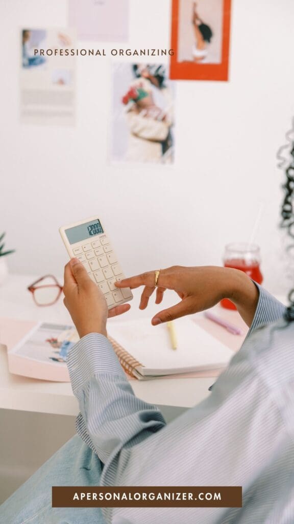 A person sits at a desk using a calculator, surrounded by notebooks, a pen, and a drink. The wall displays photos and the text “Professional Organizing” and “apersonalorganizer.com,” highlighting an organization-focused workspace.