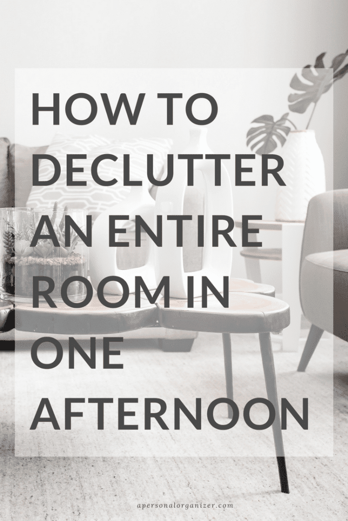 A minimalistic living room featuring a modern chair, cushions, and a potted plant in gray tones. Overlaying text reads, "how to DECLUTTER AN ENTIRE ROOM IN ONE AFTERNOON" with a small website URL at the bottom.