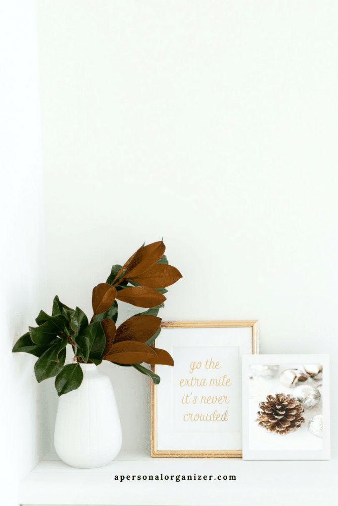 A white shelf holds a white vase with green leaves and two framed pictures. One golden frame reads "go the extra mile, it's never crowded," and the other, smaller silver frame shows a close-up of a pinecone with holiday decorations—perfect for decorating small spaces. Website "apersonalorganizer.com" is at the bottom.
