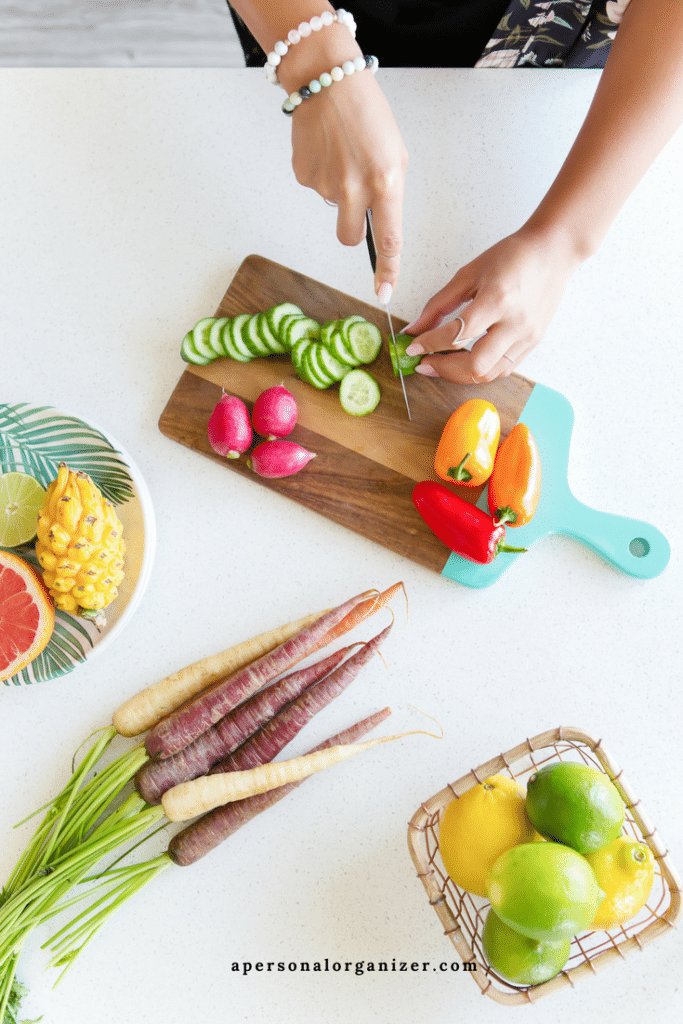 A person is slicing cucumbers on a wooden cutting board with colorful radishes and peppers beside it while planning their menu. A basket of lemons and limes, heirloom carrots, and a plate with pineapple and grapefruit are arranged on the counter, alongside the website "apersonalorganizer.com" visible.