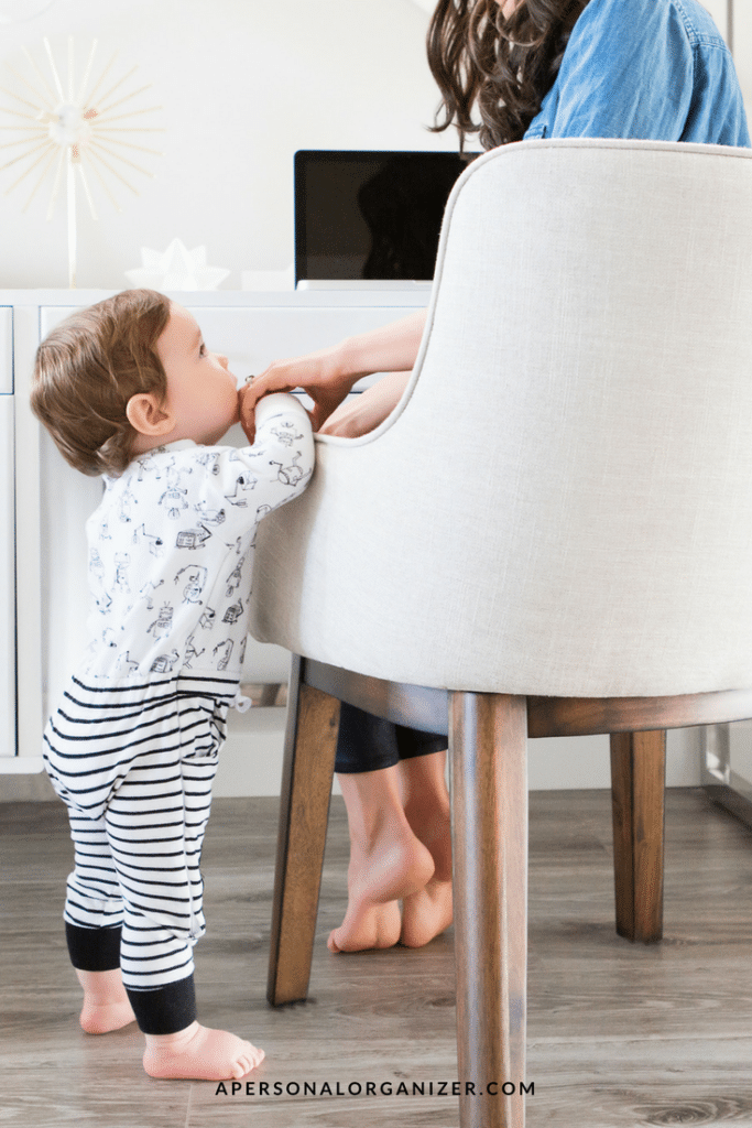 A toddler in striped pants and a patterned top stands and leans on a white cushioned chair where an adult is seated, working on a laptop. The setting, with its wooden flooring and bright indoor space, subtly captures the essence of home work balance. The adult's face remains out of view.