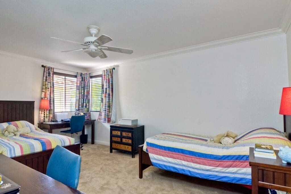 A bedroom with two single beds, each with a striped bedspread. The room has a ceiling fan, a window with colorful curtains, and a small desk with a blue chair. A chest of drawers and a bedside table with a red lamp complete the scene. Stuffed toys on each bed add charm to this post-military transition space.