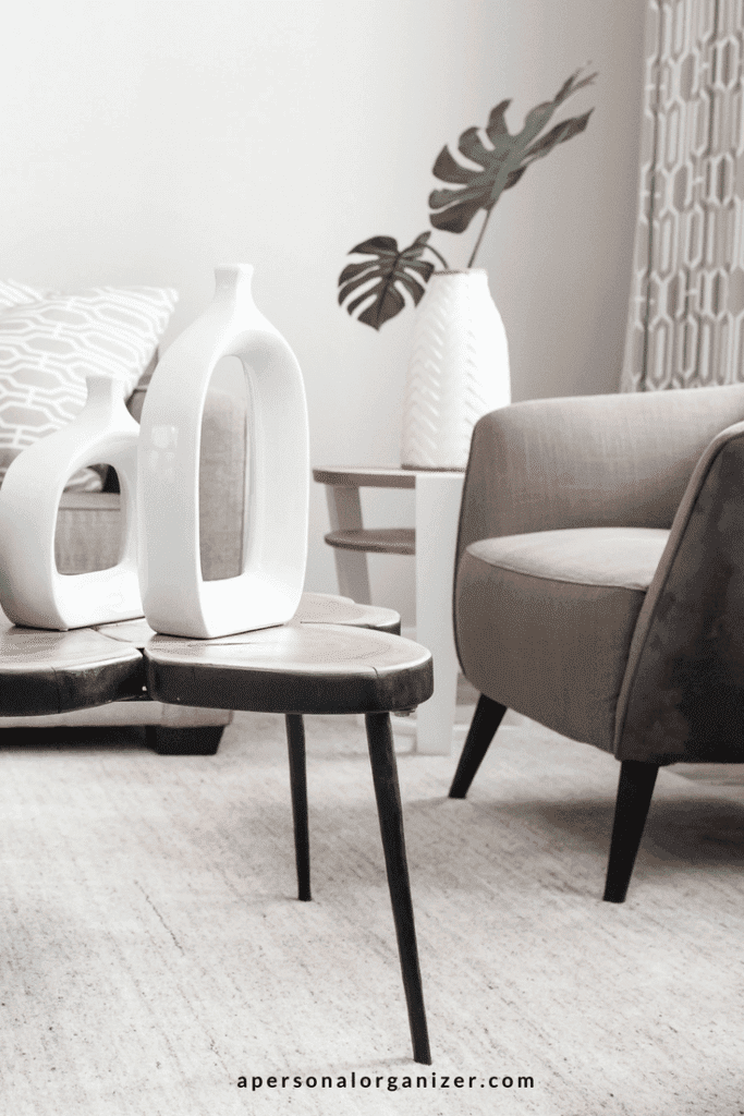 A modern living room features a decluttered space with a gray armchair, a decorative side table holding a white vase and plant, and a coffee table adorned with two abstract white vases. A gray-patterned curtain is partially visible in the background, complementing the minimalist, neutral-toned decor.