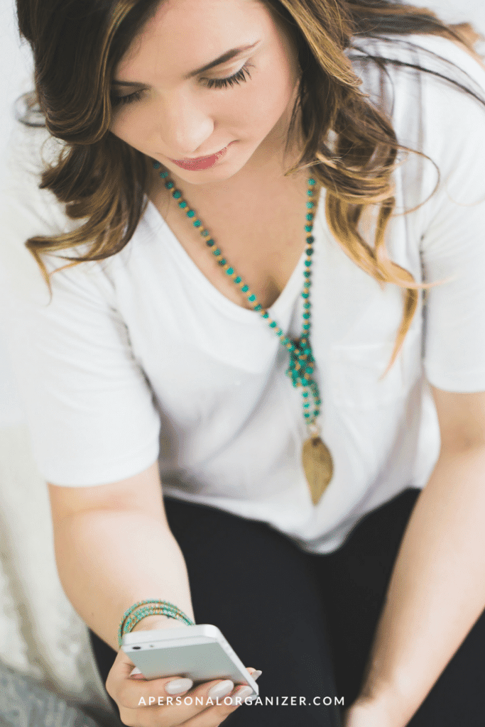 A woman with wavy brown hair wears a white top and turquoise jewelry, including a long necklace with a gold pendant. She is looking at her smartphone, which she holds in her right hand. The text "APERSONALORGANIZER.COM," an expert in nichos na organização profissional, appears at the bottom of the image.