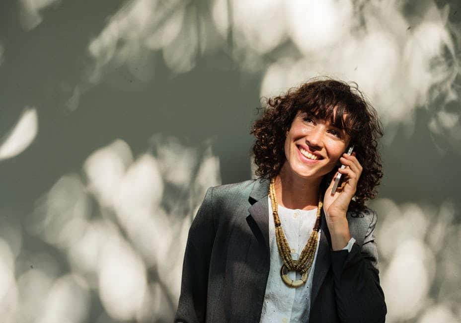A woman with curly hair, wearing a grey blazer and layered gold necklaces, smiles while talking on a cell phone. She stands against a light-colored background with dappled sunlight and shadows, embodying one of the many nichos na organização profissional that celebrate confident communication.