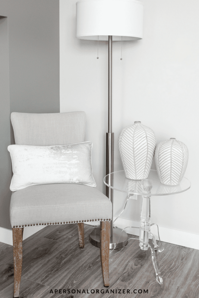 A modern interior corner showcases a light grey upholstered chair with a metallic base pillow, a tall standing lamp with a white shade, and a clear acrylic side table holding two white, patterned vases. The well-organized setup on the wooden floor against the neutral wall backdrop promotes limpeza da casa.
