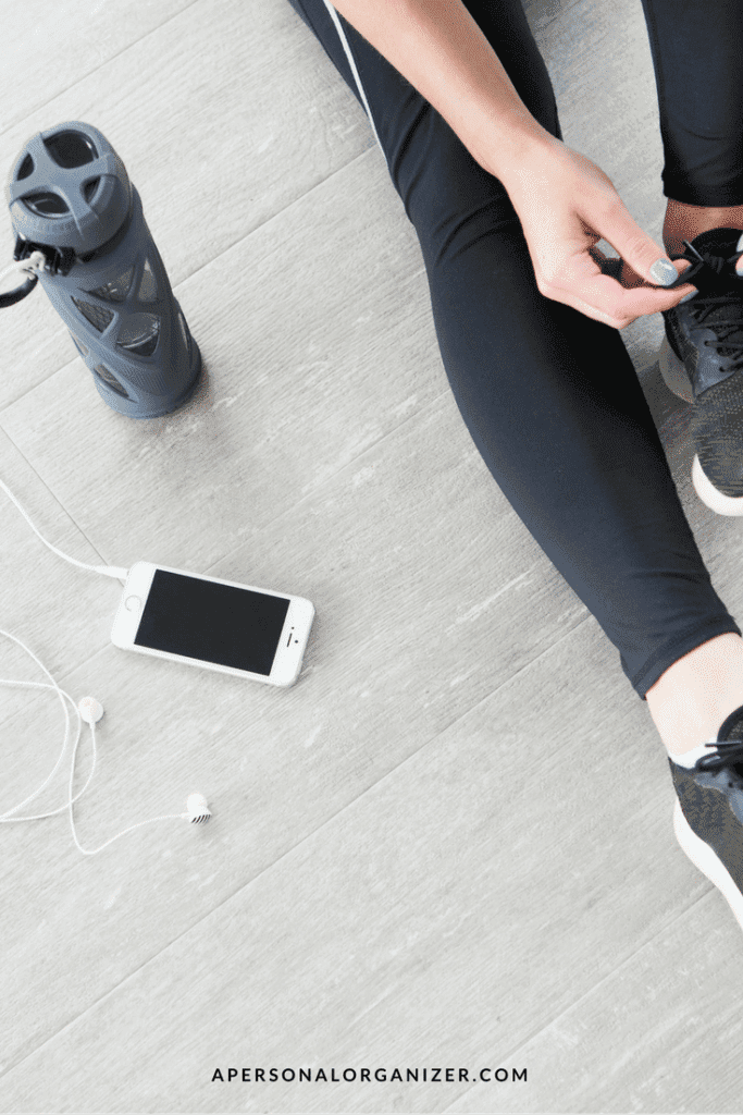 A person wearing black athletic wear ties the laces of their black sneakers. Nearby, a smartphone with earphones plugged in, and a water bottle with a handle cap are on the floor—a scene reflecting their new year's resolution. The text "APERSONALORGANIZER.COM" is visible at the bottom.