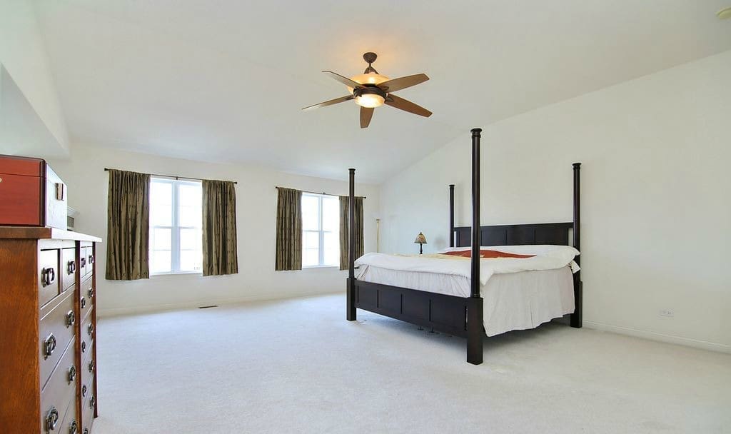 Spacious, bright bedroom with white walls and carpet, a dark wood four-poster bed with white bedding, a wooden dresser, and two large windows with green curtains—a must-see on any home tour. A ceiling fan hangs above the bed.
