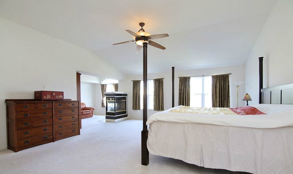 Spacious, bright bedroom featured in our home tour with a four-poster bed, wooden dresser, large windows with drapes, ceiling fan, and a cozy sitting area with fireplace and brown armchair. Walls and carpet are white.