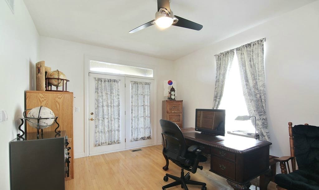 A bright home office featured in a recent home tour, with a large wooden desk, computer monitor, black office chair, bookshelves, and globes. French doors and windows with sheer curtains fill the space with natural light—a ceiling fan overhead.