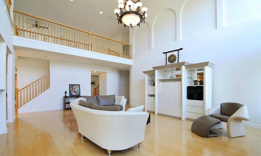 Step inside this stunning home tour: a modern living room with light wood floors, white walls, a curved white sofa, gray and brown chairs, built-in shelves, and a loft with wooden railing beneath a grand chandelier.