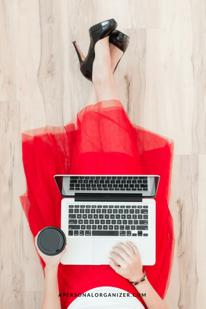 A top-down view of a person wearing a red dress and black high heels, sitting on a light wood floor while working on a silver laptop. The person holds a black cup in their left hand. The website address "APERSONALORGANIZER.COM" is at the bottom of the image, creating a Pinterest-perfect scene.