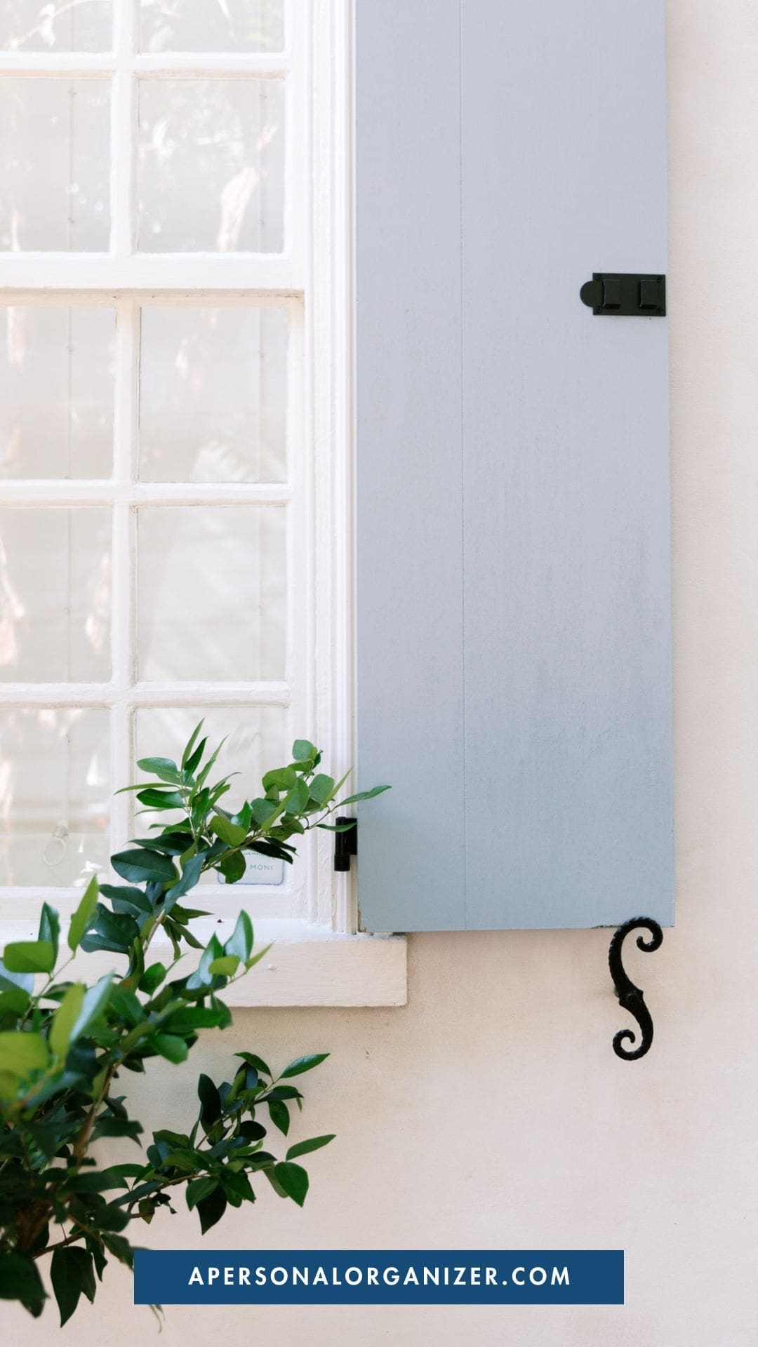 A white window with a closed pale blue shutter, black metal latch, and a curling shutter holder next to green leaves suggests serene organization; apersonalorganizer.com appears on a blue banner at the bottom.