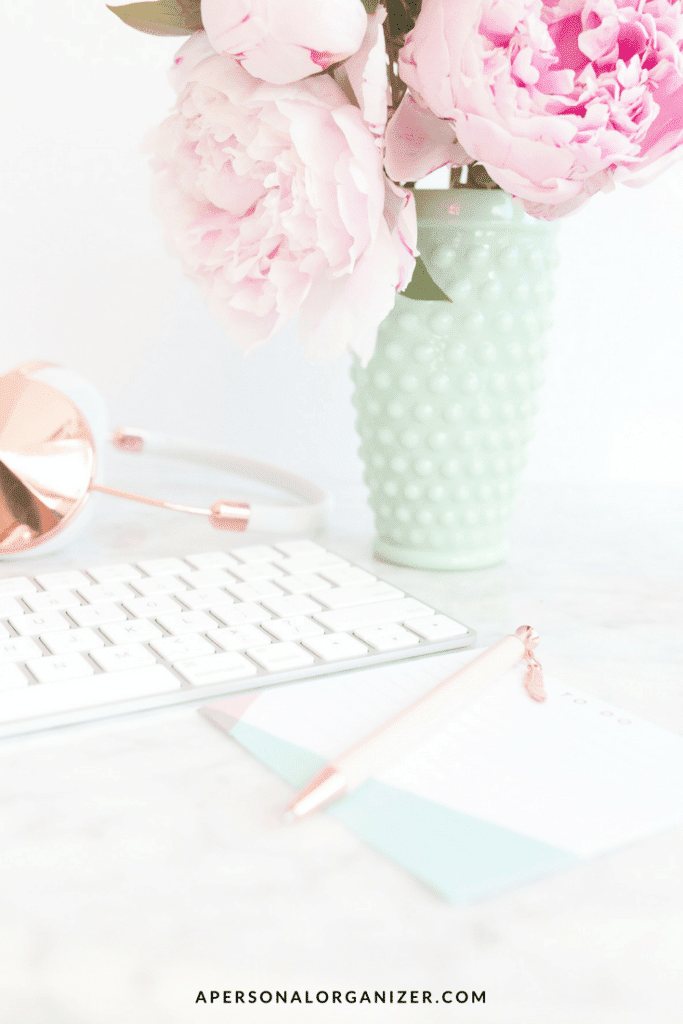A workspace featuring a white keyboard, a notepad with a pen, a pair of headphones, and a green vase filled with pink peonies. The background is light and airy. "Stay Organized" is the focus here, showcased prominently on apersonalorganizer.com at the bottom of the image.