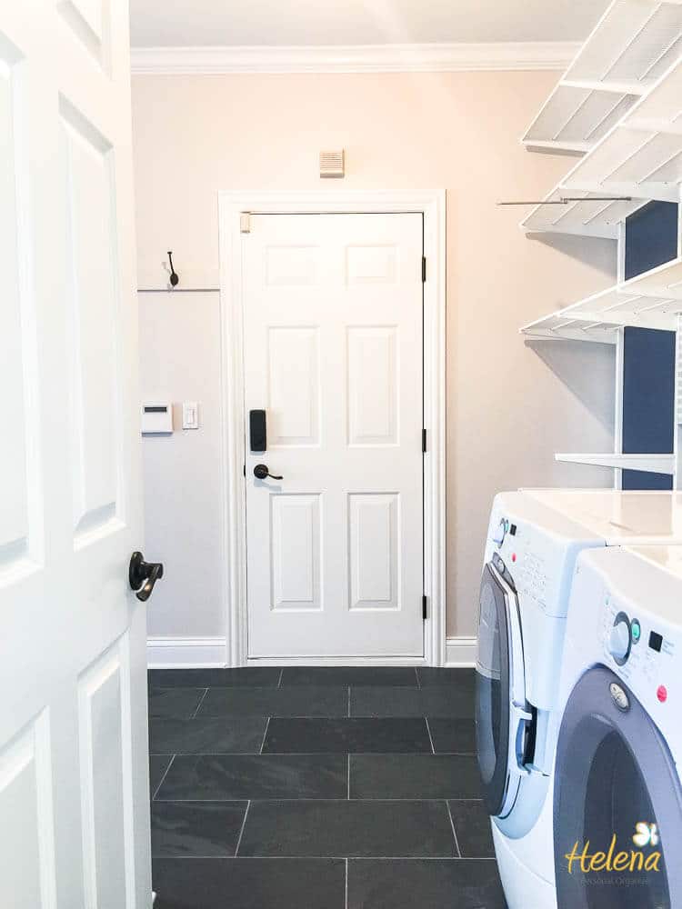 A laundry room with a white washer and dryer on the right. The space features dark tile flooring, white shelves on the walls, a white door with black hardware, and a hook on the wall. The name "Helena" is written in yellow at the bottom right.