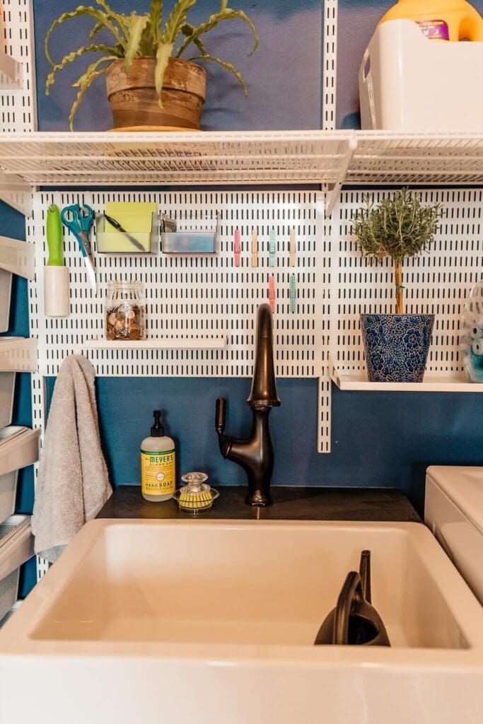 A laundry room sink with a black faucet is in front of a dark blue wall. Above the sink, shelves and a pegboard hold various items, including cleaning supplies, a potted plant, and scissors. A small towel hangs to the left of this well-organized laundry room setup.