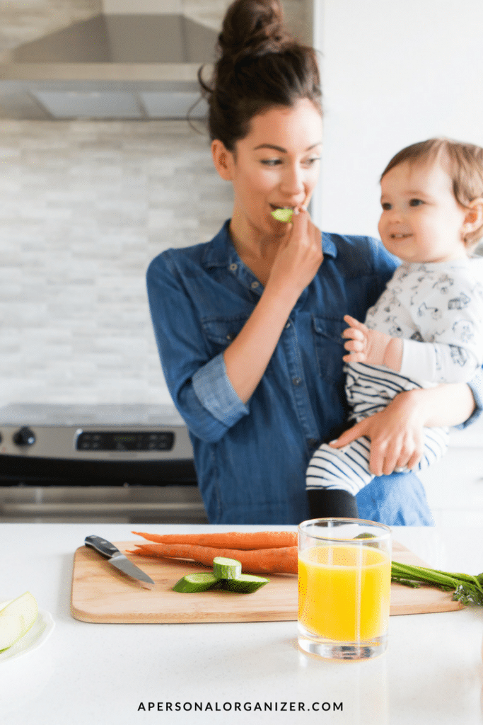 A woman stands in a kitchen holding a toddler, embodying work-life balance. She is eating a slice of cucumber and wearing a blue denim shirt. The toddler is in pajamas with a black and white pattern. On the counter are a glass of orange juice, a knife, carrot sticks, and cucumber slices.