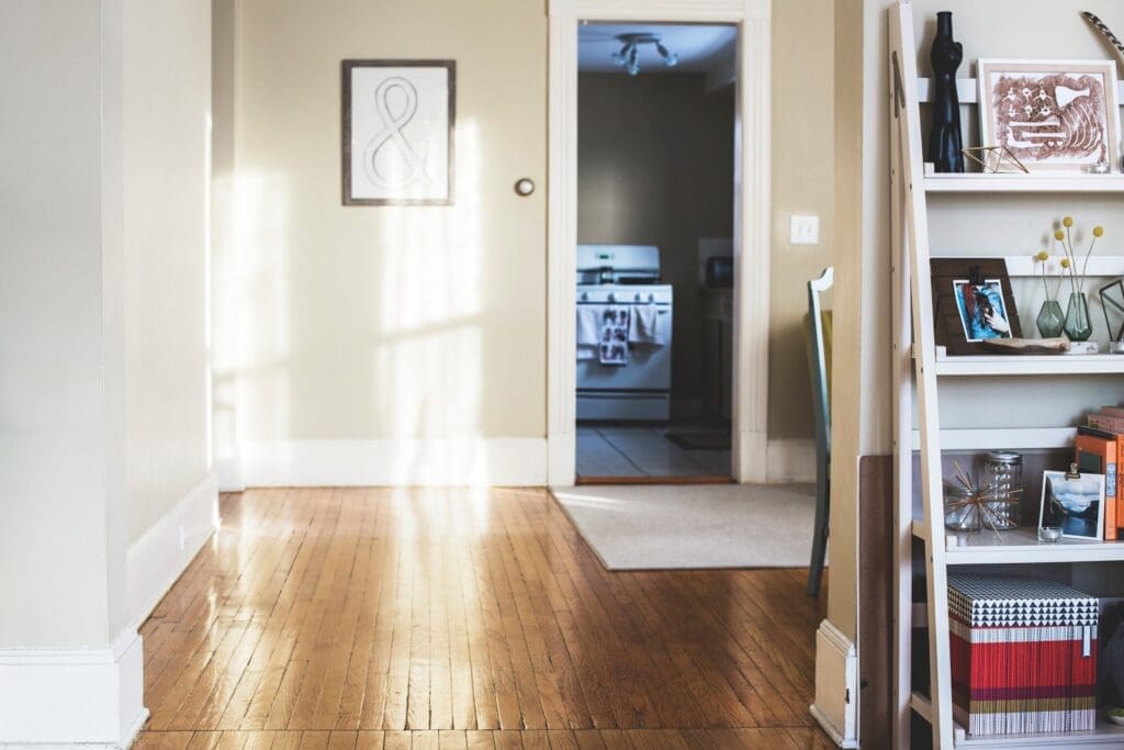 A cozy, sunlit hallway with wooden floors leads to a kitchen. On the right, a white bookshelf holds various items including framed photos, books, and decor pieces. A framed artwork hangs on the beige wall, and natural light filters through windows, adding a touch of interior design elegance.